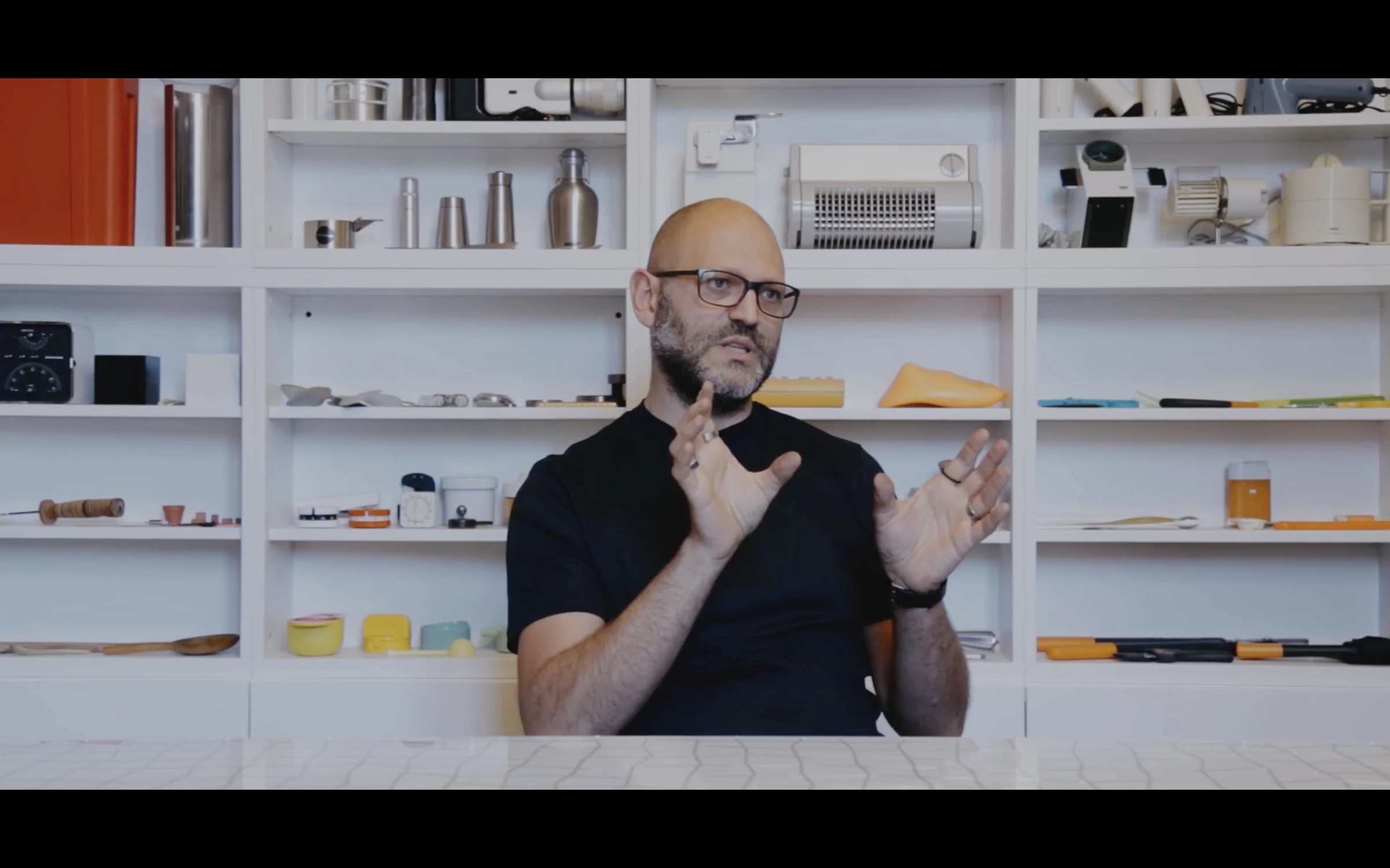 A designer gesturing mid-interview in a studio lined with shelves of models